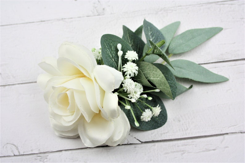 May include: A single white rose with small white flowers and green leaves. The bouquet is arranged on a white wooden surface.