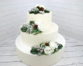 May include: A three-tiered white wedding cake decorated with white and green floral arrangements and pine cones.