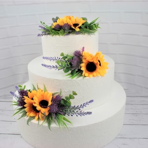 May include: A three-tiered white wedding cake decorated with artificial sunflowers, lavender, and greenery.