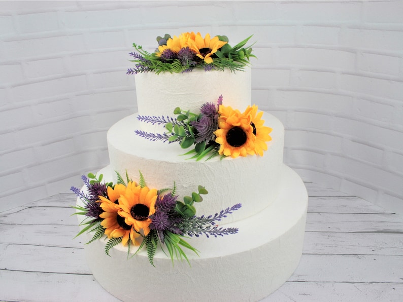 May include: A three-tiered white wedding cake decorated with artificial sunflowers, lavender, and greenery.