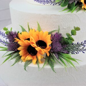 May include: A white wedding cake decorated with a floral arrangement of yellow sunflowers, purple thistle, and lavender sprigs.