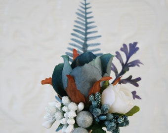 Puede incluir: Un boutonniere floral azul y blanco con un helecho, una rosa blanca y detalles plateados.