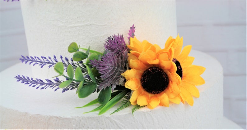 May include: A wedding cake topper featuring a bouquet of yellow sunflowers, purple thistle, lavender sprigs, and green foliage.