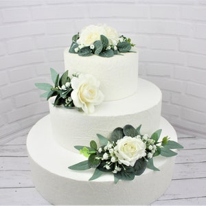 May include: A three-tiered white wedding cake decorated with white roses and greenery.