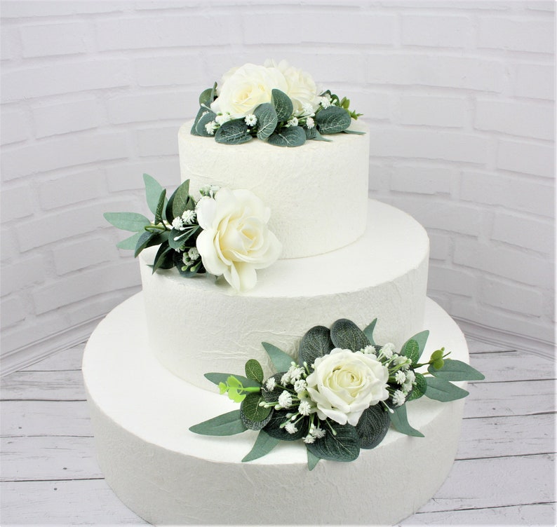 May include: A three-tiered white wedding cake decorated with white roses and greenery.