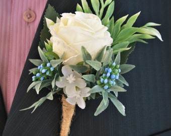 May include: A white rose boutonniere with blue and white flowers and greenery, tied with twine.