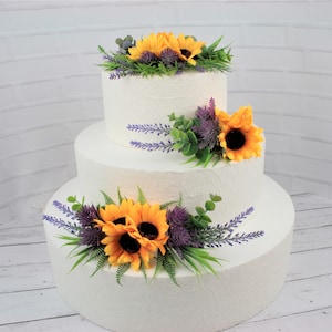 May include: A three-tiered white wedding cake decorated with yellow sunflowers, purple thistle, and lavender sprigs.