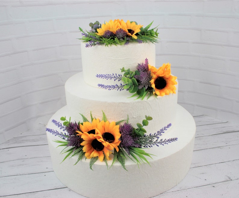 May include: A three-tiered white wedding cake decorated with yellow sunflowers, purple thistle, and lavender sprigs.