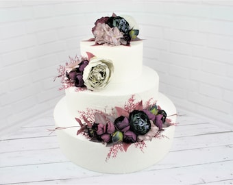 May include: A three-tiered white cake decorated with dark purple, green, and light pink flowers. The cake is adorned with floral arrangements on each tier, creating a visually appealing centerpiece. The cake sits on a white wooden surface.