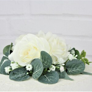 May include: White rose and greenery wedding cake topper. The topper features two white roses with green leaves and small white flowers.