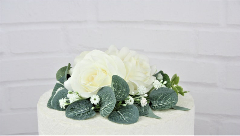 May include: White rose and greenery wedding cake topper. The topper features two white roses with green leaves and small white flowers.