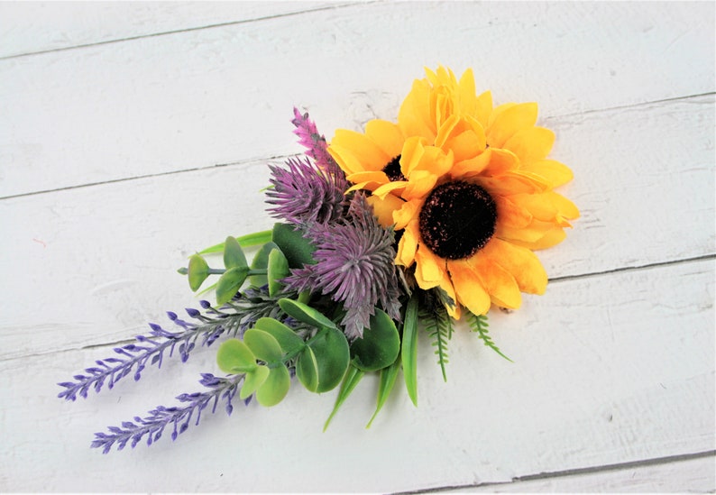 May include: A bouquet of artificial flowers featuring a yellow sunflower, purple thistle, and lavender sprigs with green eucalyptus leaves.