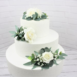 May include: A three-tiered white wedding cake decorated with white roses and greenery.