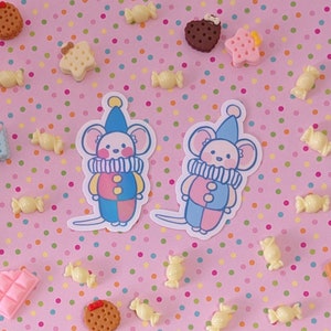 May include: Two colorful mouse stickers with clown costumes. The mice are wearing red, blue, and pink. The stickers are on a pink background with white polka dots.