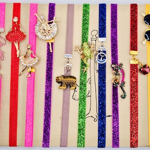 May include: Ten colorful glittery bookmarks with various charms, including a ballerina, a bear, a frog, a lizard, and a heart. The bookmarks are on a white background.