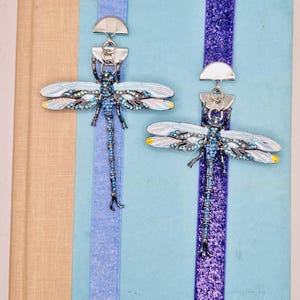 May include: Two dragonfly bookmarks with blue and white wings, embellished with rhinestones, hang from a light blue book. The bookmarks have silver-colored metal accents and are set against a light blue and tan background.