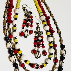 May include: A multi-strand necklace with red, white, yellow, and black beads. The necklace is made with a variety of materials, including glass, wood, and metal. The necklace is paired with matching earrings.