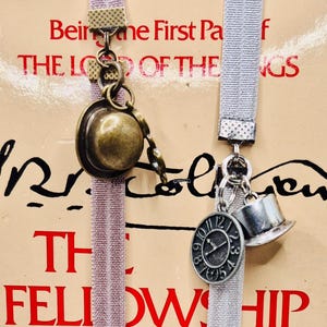 Può includere: Due segnalibri decorativi in nastro con ciondoli in metallo. Uno ha un ciondolo a forma di cappello color bronzo, l'altro un orologio e un cappello a cilindro. Il testo del libro recita "Being the First Part of The Lord of the Rings" e "The Fellowship".