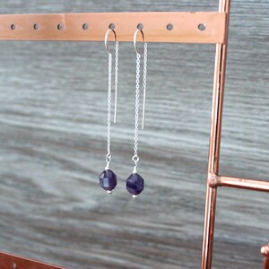 May include: A pair of silver threader earrings with faceted, deep purple amethyst beads. The earrings hang from a copper-colored jewelry display stand against a gray wooden background. The earrings are delicate and elegant.