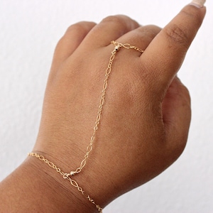 May include: A delicate gold hand chain bracelet. The jewelry piece connects a ring to a bracelet with a fine chain. The ring and bracelet feature small, round beads. The hand chain is worn on a hand with a light skin tone.