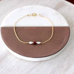 May include: A delicate gold chain bracelet featuring a central arrangement of two small white oval pearls flanking a single deep red round bead. The bracelet includes a gold lobster claw clasp and is designed with a thin, polished metal link chain.
