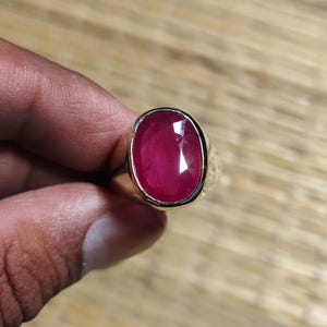 May include: A gold ring featuring a large, oval-cut, deep pinkish-red gemstone set in a smooth, polished metal bezel. The stone displays multiple facets that catch the light, creating a vibrant, saturated color profile within the gold band.
