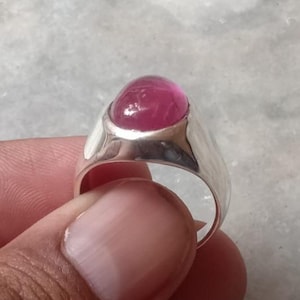 Może przedstawiać: Srebrny pierścionek z dużym, owalnym, jasnoróżowym kamieniem szlachetnym. Pierścionek ma prostą, wypolerowaną obrączkę i oprawę w kształcie ramki, która bezpiecznie trzyma kamień szlachetny. Kolor kamienia szlachetnego jest głęboki, nasycony różem.