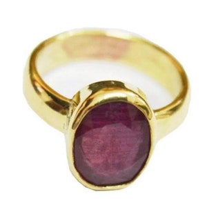 May include: A gold ring featuring a large, oval-shaped ruby gemstone. The ruby is a deep red colour and has a smooth, polished surface.