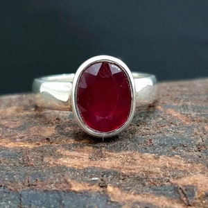 May include: A silver ring featuring a large, oval-shaped ruby gemstone. The ruby is a deep red colour and has a faceted cut.