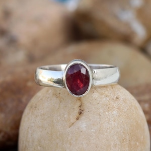 May include: A silver ring with a red oval gemstone. The ring is set on a light brown stone.