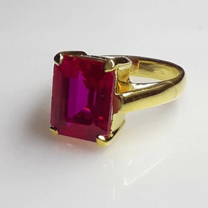 Könnte beinhalten: Ein goldener Ring mit einem großen, smaragdgeschliffenen, rubinroten Edelstein.