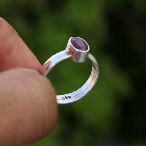 Pode incluir: Um anel de prata com uma pequena gema ovalada de cor rosa escura. O anel &eacute; marcado com "925" na banda interior.