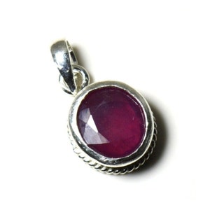 May include: A silver pendant with a deep red oval ruby gemstone. The pendant has a detailed border around the stone.