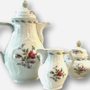 May include: A white porcelain tea set with a floral design. The set includes a teapot, creamer, sugar bowl, and coffee pot. Each piece is decorated with pink roses and green leaves.