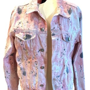 Op de afbeelding: Een roze denim jack met een splatter paint design in zwart en grijs.