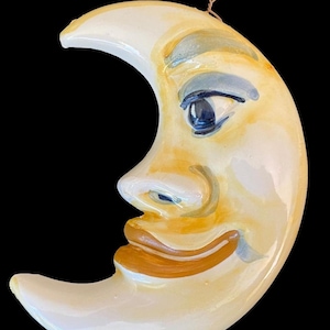 May include: A ceramic crescent moon ornament with a painted face. The moon is yellow with a white outline and has a blue eye, brown mouth, and gray eyebrow.