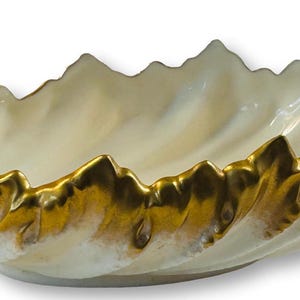 May include: An off-white ceramic bowl with a gold-coloured rim. The rim has a mountain-like design. The bowl has a swirl detail on one end. The bowl is approximately 30 cm long.