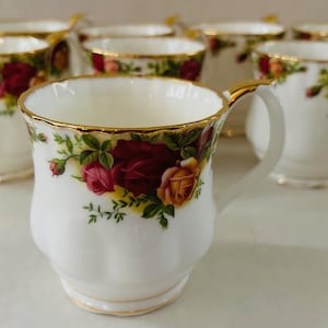以下が含まれることがあります： 金色の縁と取っ手が付いた、白い磁器製のティーカップ8個セット。各カップは、赤と黄色のバラと緑の葉のフローラルパターンで装飾されています。白い表面に配置されています。