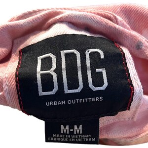Op de afbeelding: Roze tie-dye denim jack met een zwart label met de tekst "BDG Urban Outfitters" en een kleiner wit label met de tekst "M-M Gemaakt in Vietnam Fabriqu&eacute; en Vietnam".