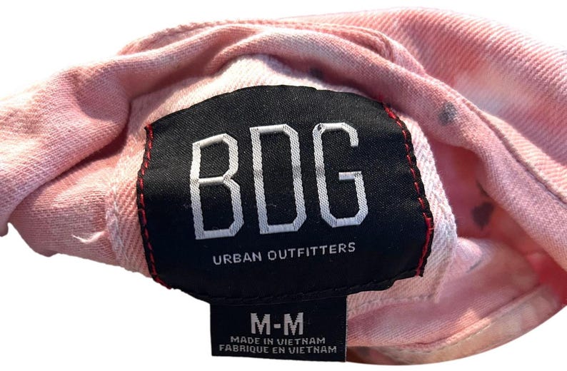 Op de afbeelding: Roze tie-dye denim jack met een zwart label met de tekst "BDG Urban Outfitters" en een kleiner wit label met de tekst "M-M Gemaakt in Vietnam Fabriqu&eacute; en Vietnam".