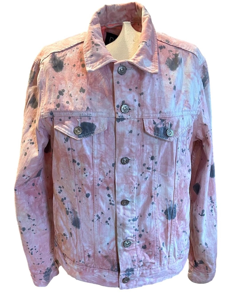 Op de afbeelding: Een roze denim jack met een tie-dye patroon en zwarte verfspatten.