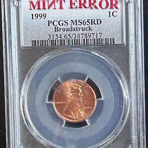 Può includere: Una moneta da un centesimo Lincoln del 1999 color rame, classificata come errore di conio broadstruck da PCGS come MS65RD, sigillata in una custodia protettiva rettangolare in plastica trasparente. L'etichetta riporta MINT ERROR, 1999, 1C, PCGS MS65RD, Broadstruck.