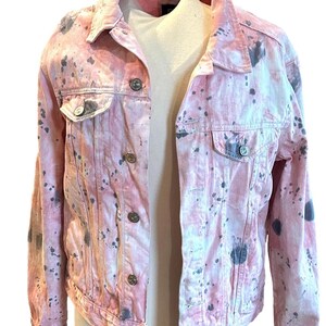 Op de afbeelding: Een roze denim jack met een splatter paint design in grijstinten. Het jack heeft een klassieke jean jacket stijl met een knoopsluiting en twee zakken.