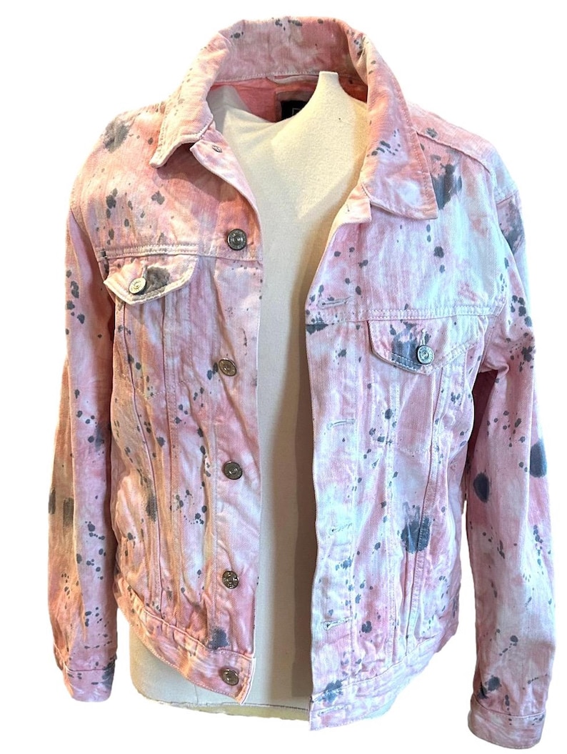 Op de afbeelding: Een roze denim jack met een splatter paint design in grijstinten. Het jack heeft een klassieke jean jacket stijl met een knoopsluiting en twee zakken.