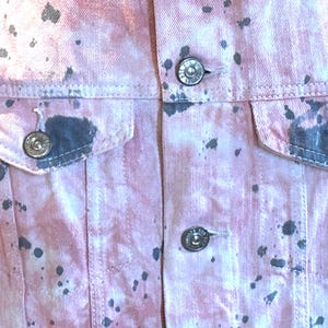 Op de afbeelding: Een roze denim jack met zwarte splatter verf. Het jack heeft twee voorzakken en een knoopsluiting.
