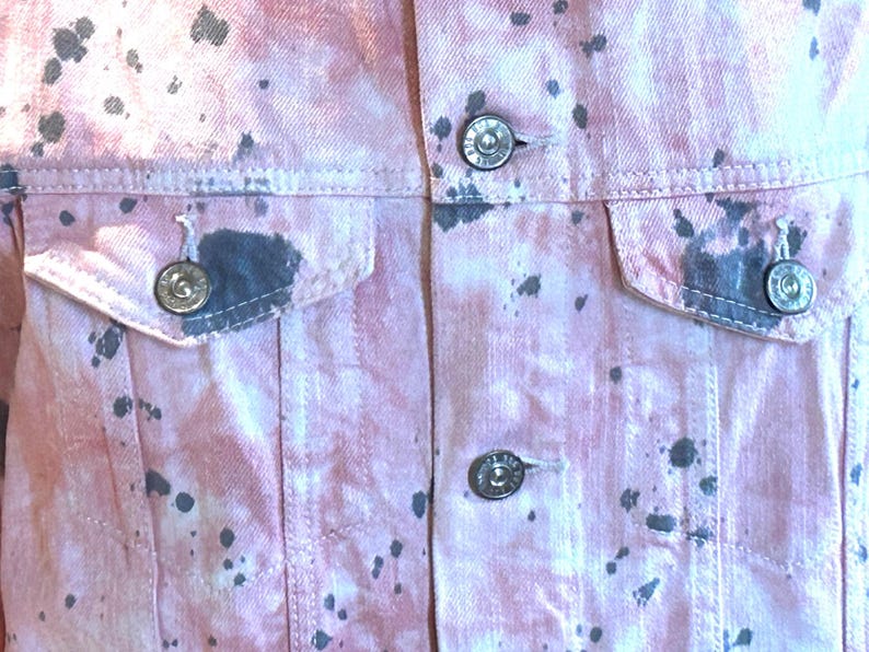 Op de afbeelding: Een roze denim jack met zwarte splatter verf. Het jack heeft twee voorzakken en een knoopsluiting.