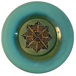 May include: A round ceramic plate with a turquoise and green glaze. The center of the plate features a starburst design in black and gold. The plate has a slightly raised edge.
