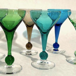May include: Six stemmed cocktail glasses with round bases. The glasses are made of clear glass and have colored stems and bases. The colors are blue, green, teal, and brown.