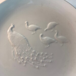 May include: A white ceramic plate with an embossed design of a peacock and four birds. The peacock has a long tail with many feathers. The birds are arranged around the peacock.