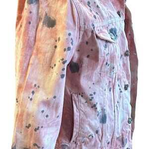 Op de afbeelding: Een roze denim jack met zwarte verfspatten. Het jack heeft een klassieke denim jack stijl met een knoopsluiting en twee zakken.